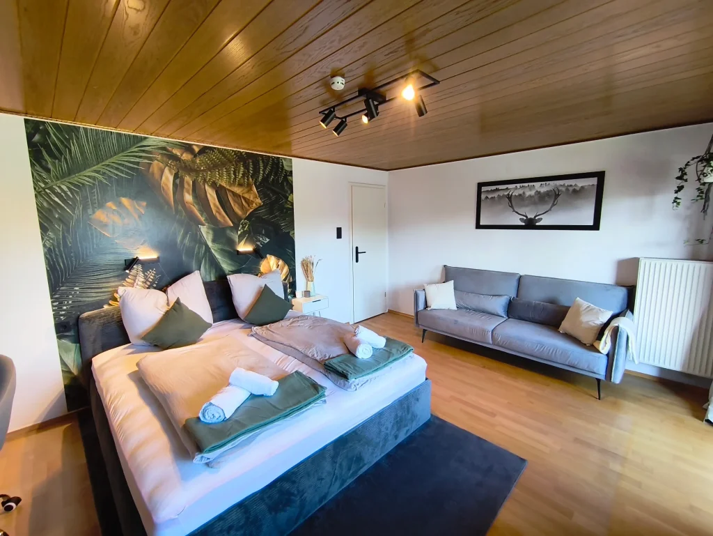 Wohn- und Schlafbereich der Ferienwohnung Burgperle mit Bett, Sofa und tropischer Fototapete