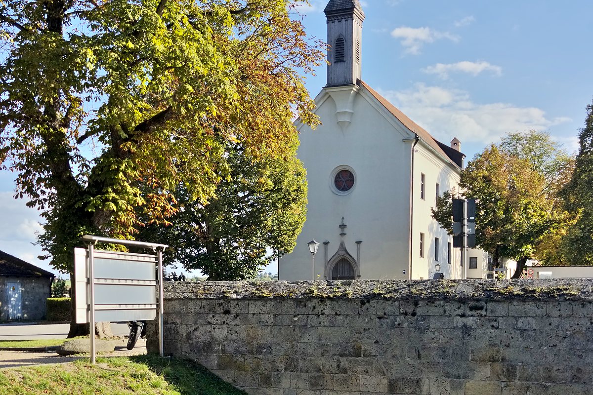 Weiße Kirche mit spitzem Turm hinter einer Natursteinmauer, davor eine Wiese