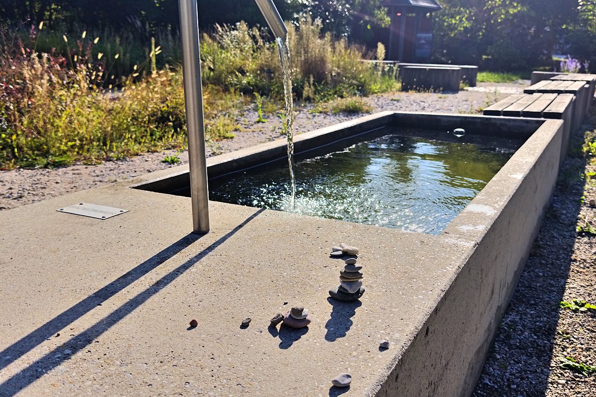 Wassertretbecken aus Beton mit Haltestange und sprudelndem Zulauf am Wegrand