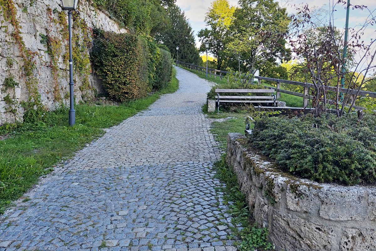 Gepflasterter Weg entlang einer hellen Felswand, mit Laterne und Bank